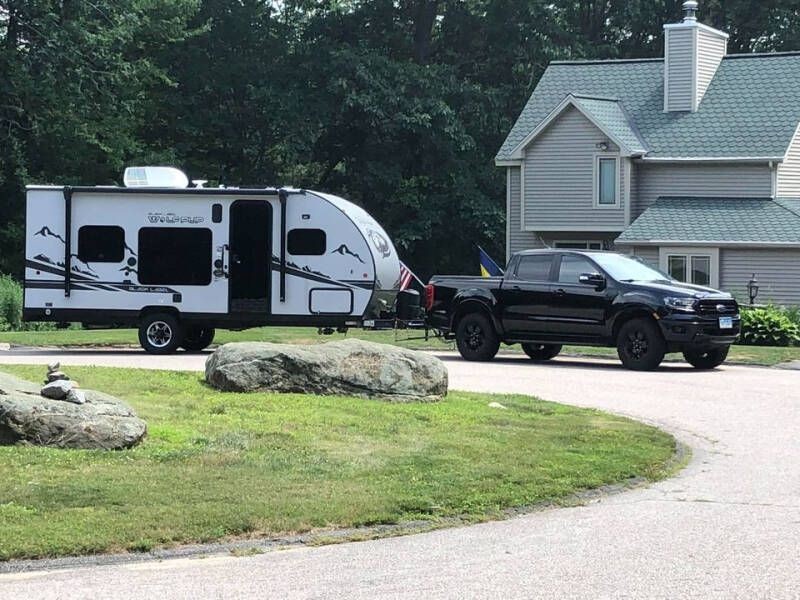 A black truck towing a white travel trailer parked in front of a suburban house with green roofing.